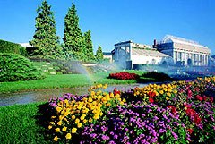 Gepflegte Gartenanlage mit bunten Blumenbeeten, Rasen und Nadelbäumen vor einem Gebäude, im Hintergrund ein Rasensprenger